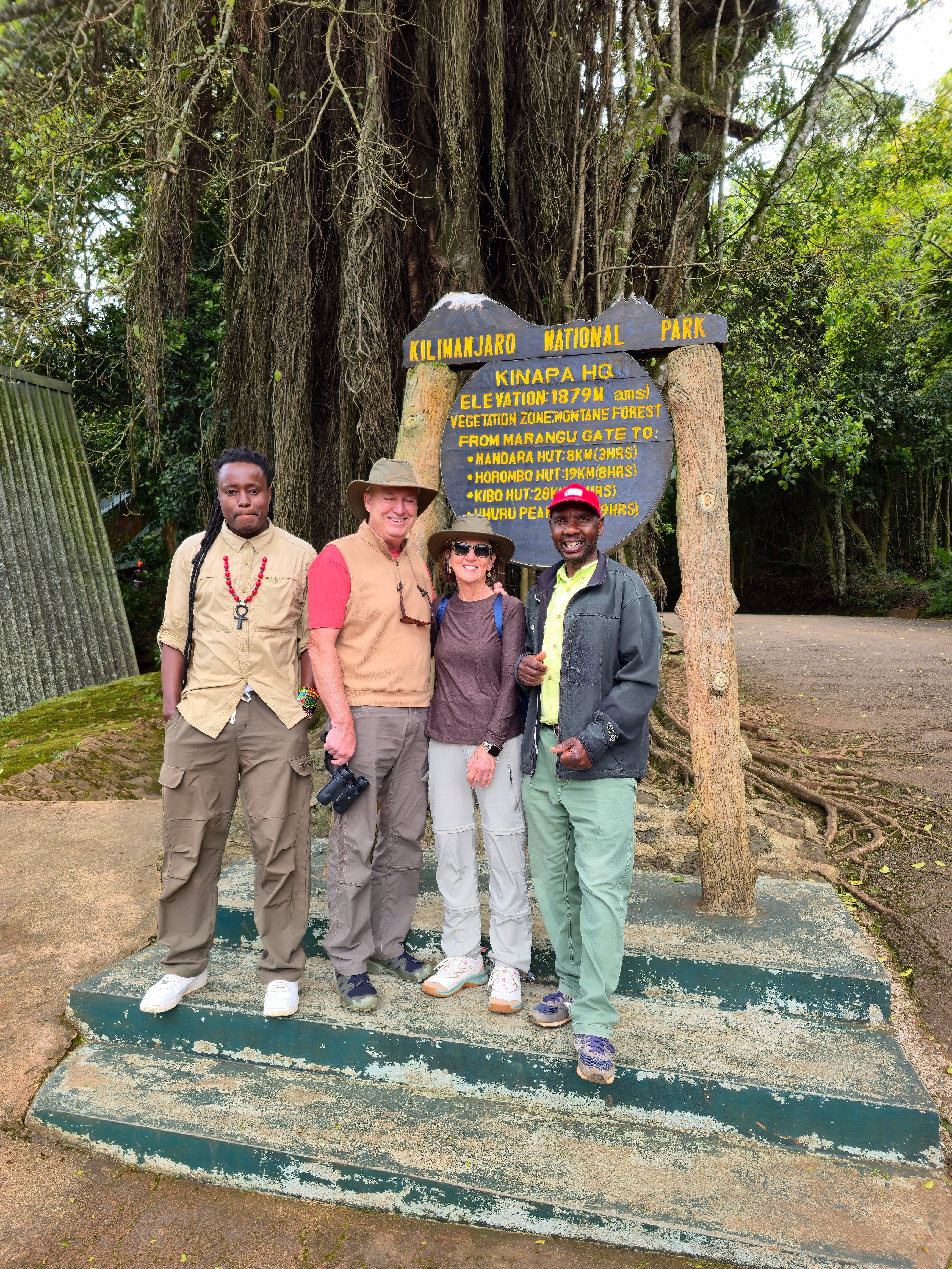 A small group of travelers posing with their guides in front of a Mount Kilimanjaro wooden park sign.