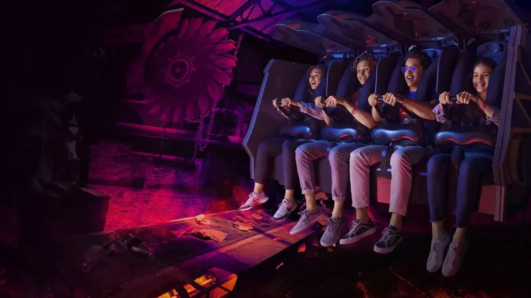 Four riders hold onto their safety restraints while seated in a suspended ride vehicle in a dark, red-lit industrial chamber at Warner Bros. World Abu Dhabi Theme Park.