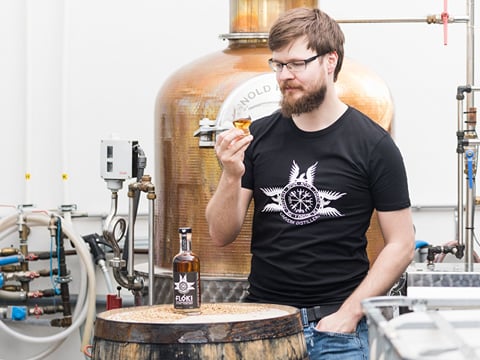 A man standing in front of a distilling vessel about to sample a spirit