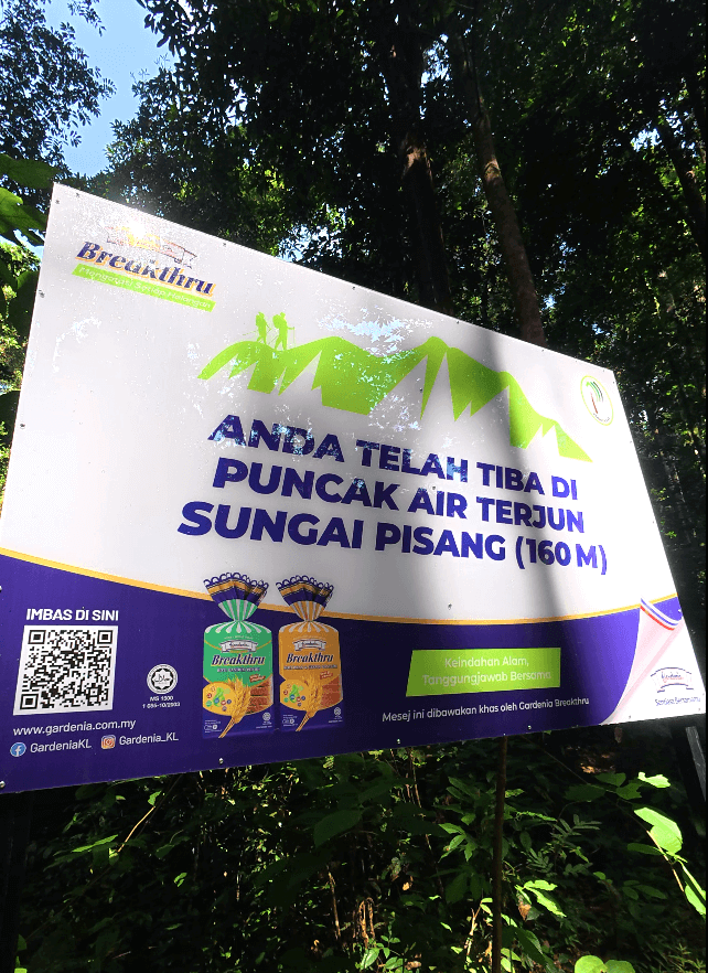 Signboard in Malay language marking the arrival at the 160-meter peak of Sungai Pisang Waterfall trail, surrounded by lush forest.