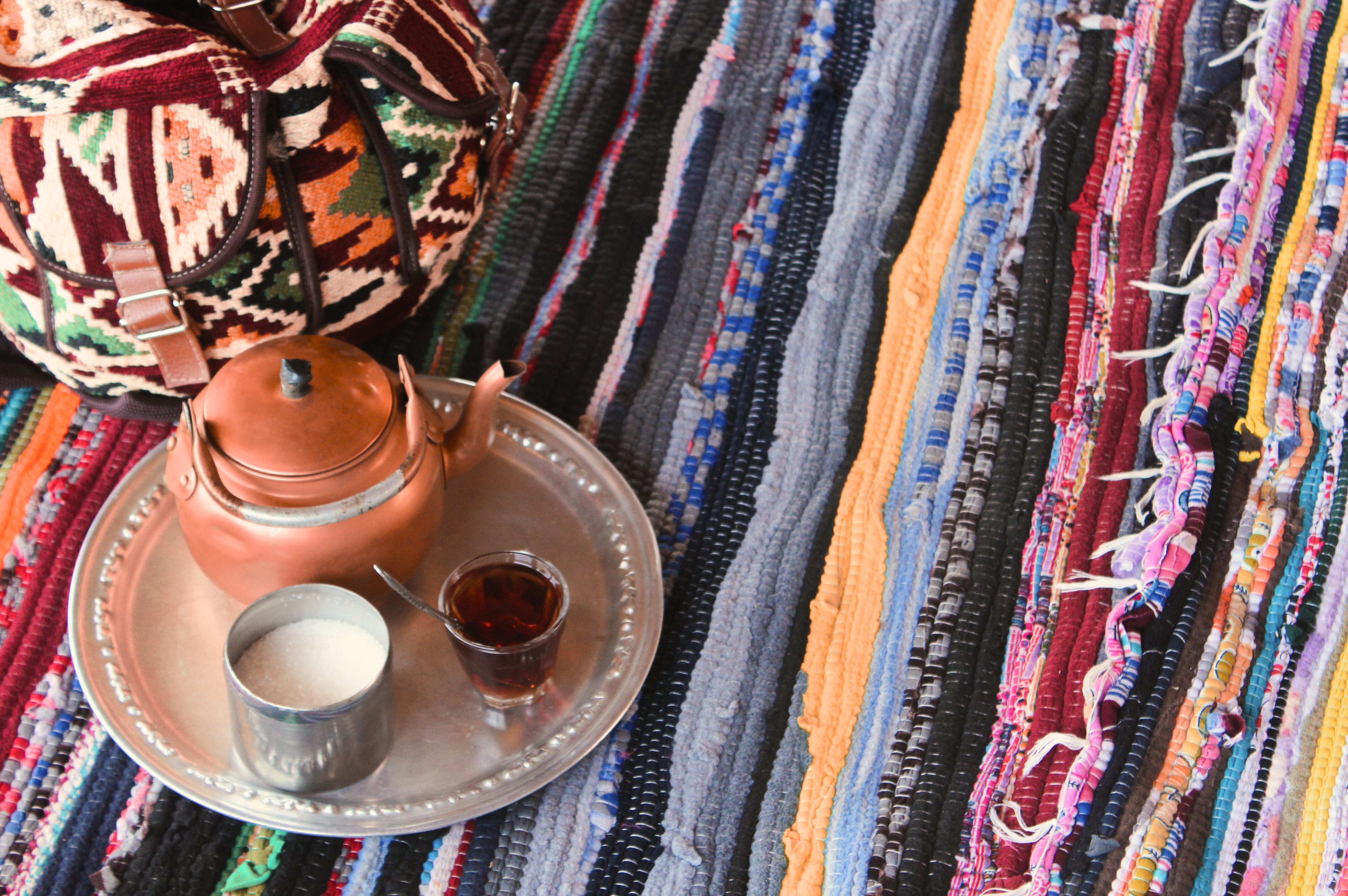 Close-up of a copper teapot, tray, and glass of tea set on a brightly colored, multicolored, textured woven desert rug.