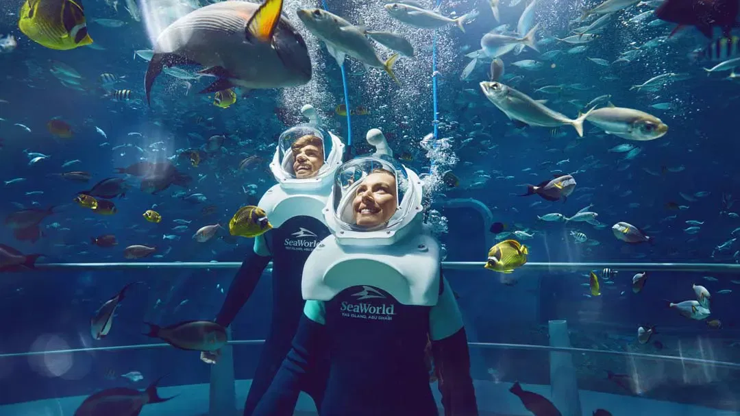 Two visitors wearing diving helmets and black wetsuits submerged underwater, surrounded by a large school of tropical fish at Sea World Abu Dhabi.