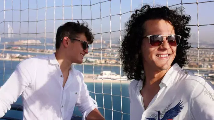 Two smiling men in white shirts and sunglasses stand against a wire mesh backdrop overlooking a coastal landscape at The Dubai Balloon at Atlantis.