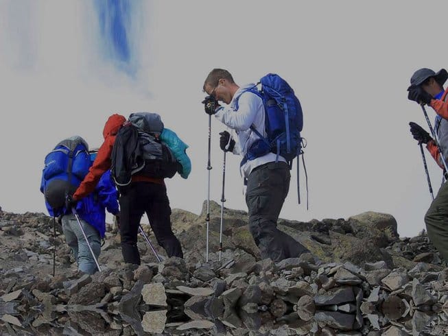 A group of trekkers wearing backpacks taking photos and walking across a rugged, rocky mountain trail.