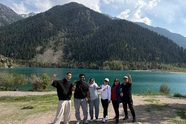 Group of tourists posing near turquoise Issyk Lake surrounded by pine-covered mountains during a jeep tour to Issyk Lake and Turgen Gorge in
