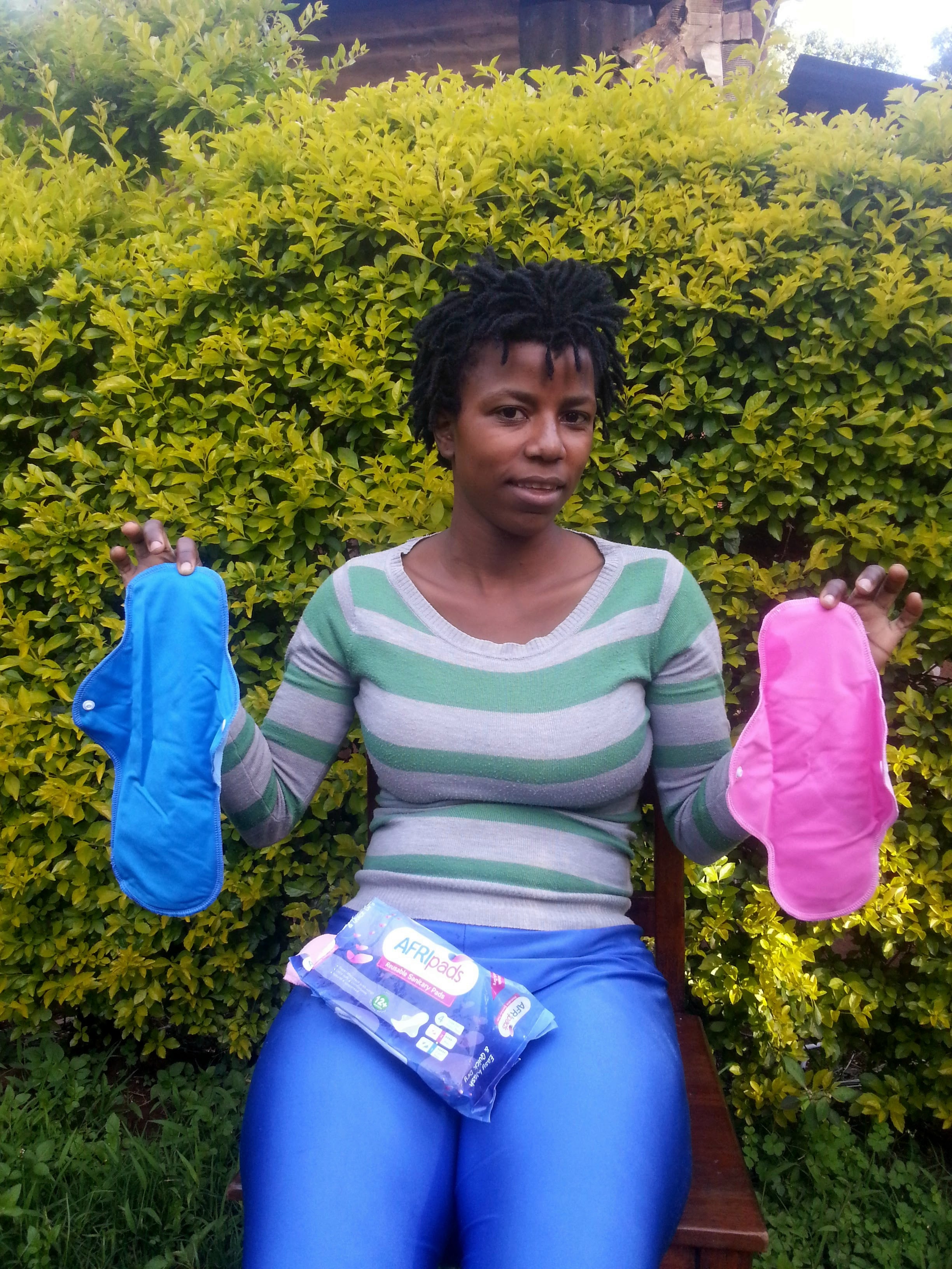A woman holding blue and pink reusable sanitary pads while sitting outdoors in front of green foliage.