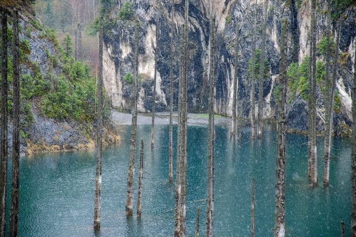 Kaindy Lake with submerged pine trees rising from turquoise water surrounded by forested mountains