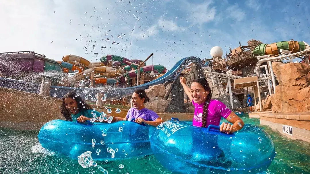 Three smiling women float happily in blue inflatable tubes amidst splashing water in front of various water slides at Yas Waterworld, Abu Dhabi.