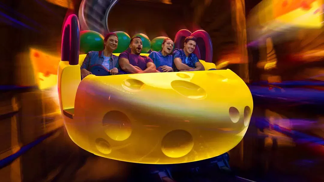 Four adults react with excitement inside a yellow cheese-shaped coaster car as it speeds through an indoor ride track at Warner Bros. World Abu Dhabi Theme Park.