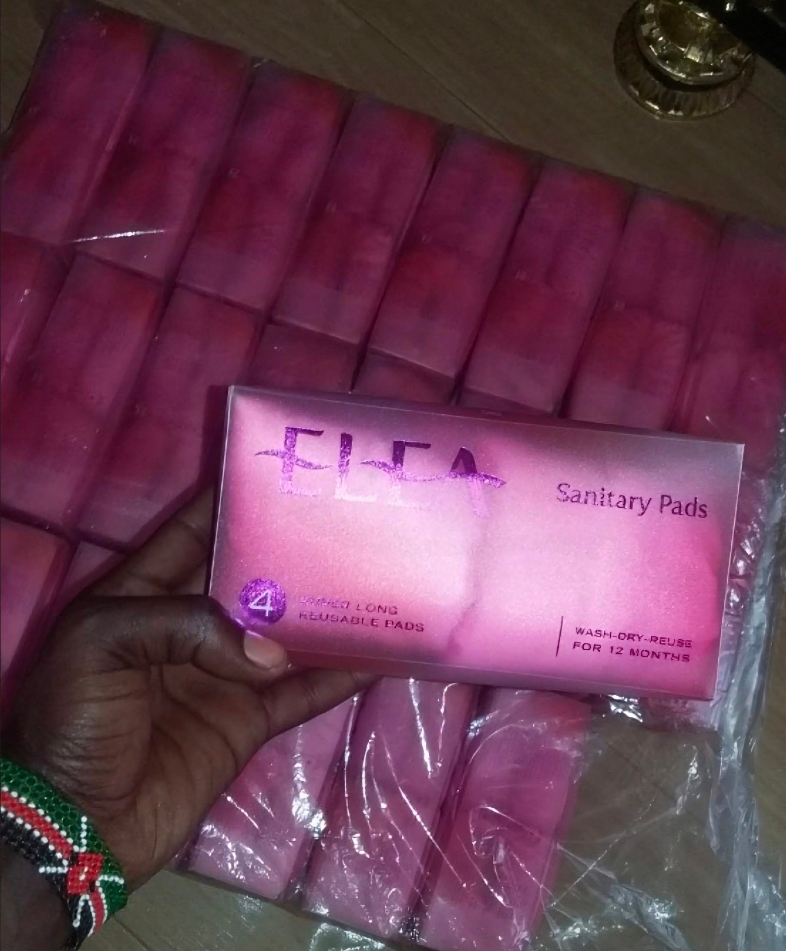 A hand holding a pack of pink ELIA reusable sanitary pads against a background of stacked pads