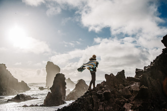 A woman draped in an icelandic woollen blanket stands on a rock on Djúpalónssandur beach taking in the views
