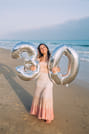 A woman stands on a sandy beach holding two large balloons shaped like the numbers "3" and "0." The ocean waves and a pale sky are visible i