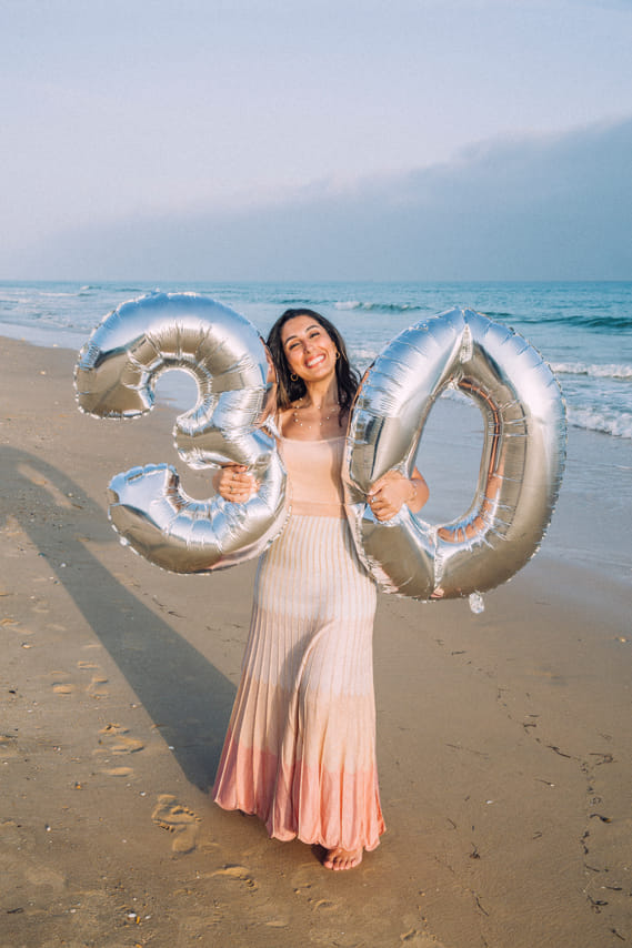 A woman stands on a sandy beach holding two large balloons shaped like the numbers "3" and "0." The ocean waves and a pale sky are visible i