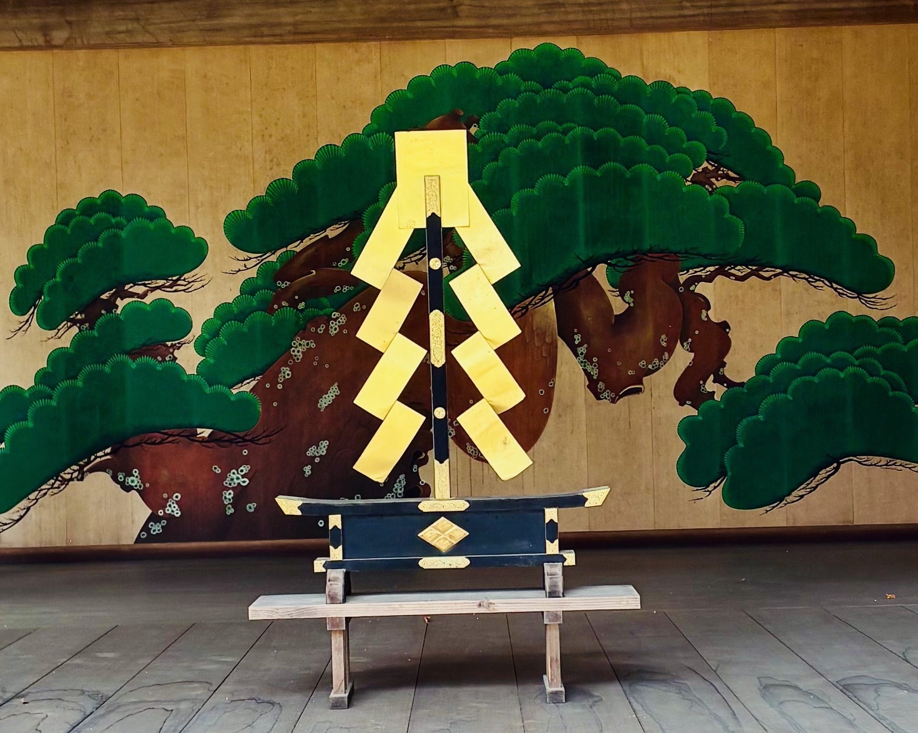 A golden decorative ornament on a wooden stand on a traditional Noh stage, in front of the pine tree mural on the back wall.