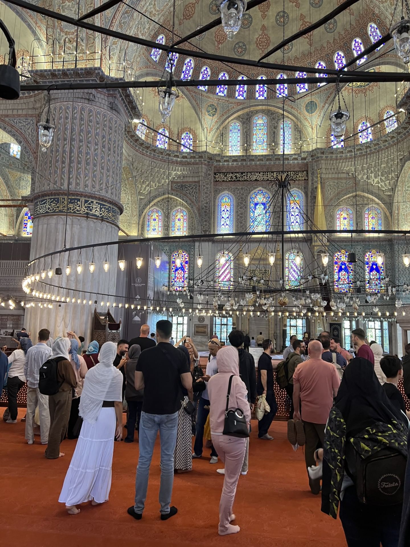 “A group of tourists inside the Blue Mosque, listening to a guide and admiring the mosque’s intricate interior decorations and architecture.