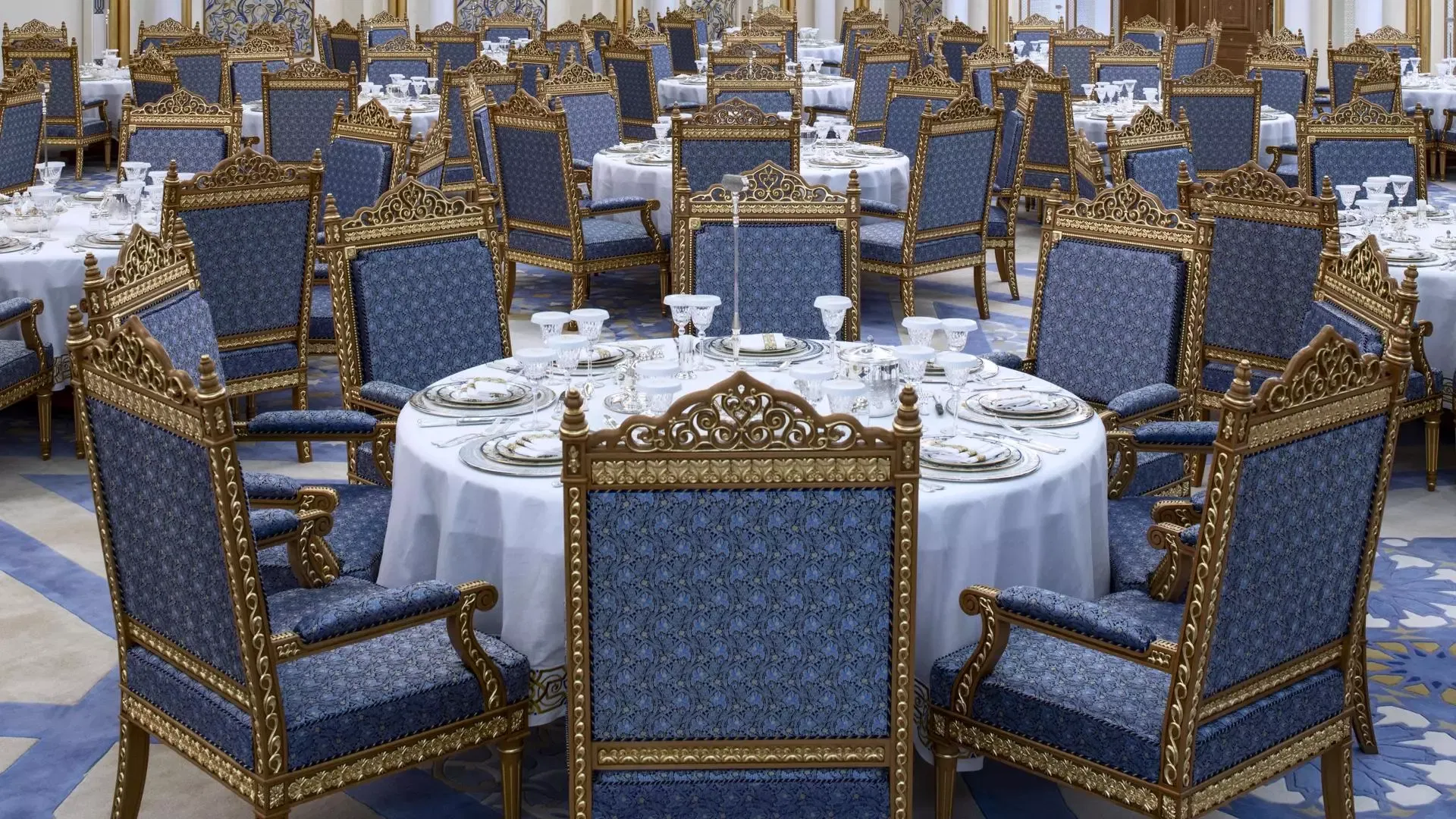 Multiple round dining tables are surrounded by tall, ornate blue and gold chairs in a grand banquet hall at Qasr Al Watan, Royal Palace.