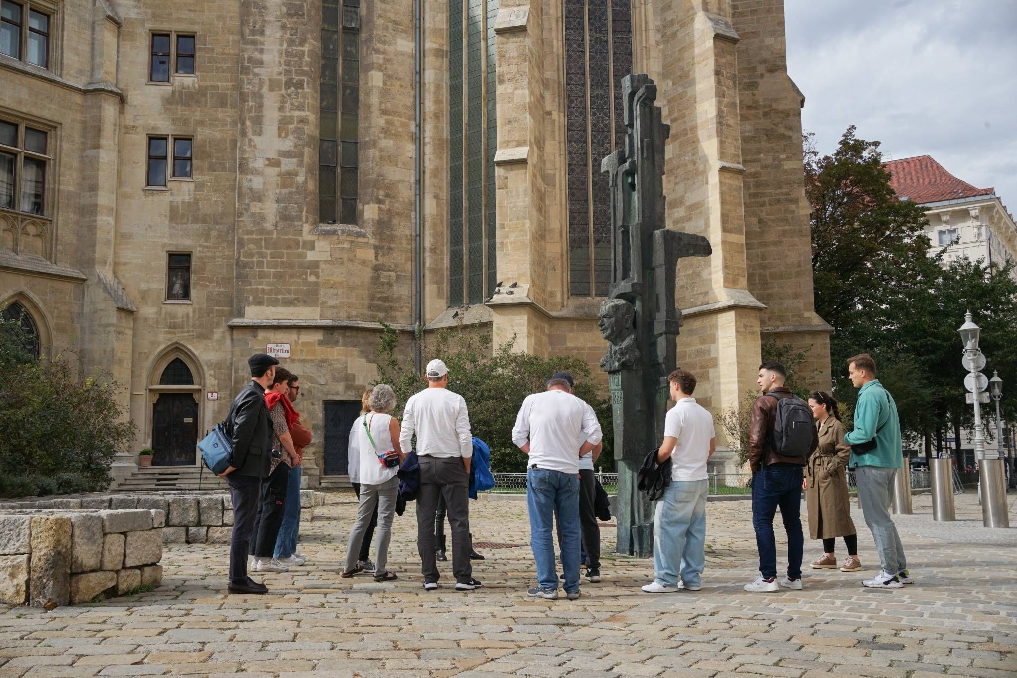 People standing in a circle near a bronze statue, with tall arched windows of a medieval stone church in the background
