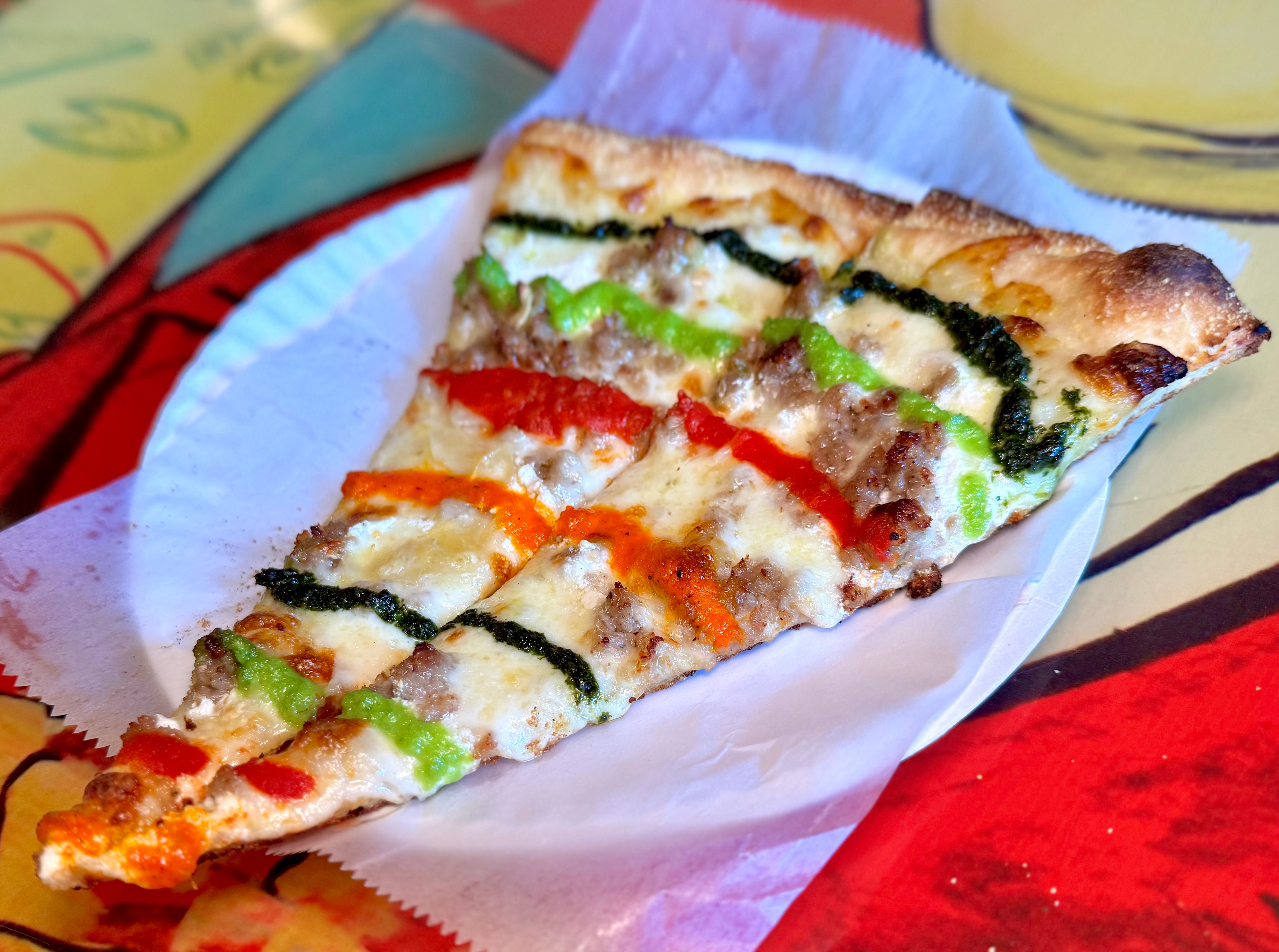 A triangular pizza slice topped with cheese, herbs, vegetables, and colorful sauces on a paper plate.