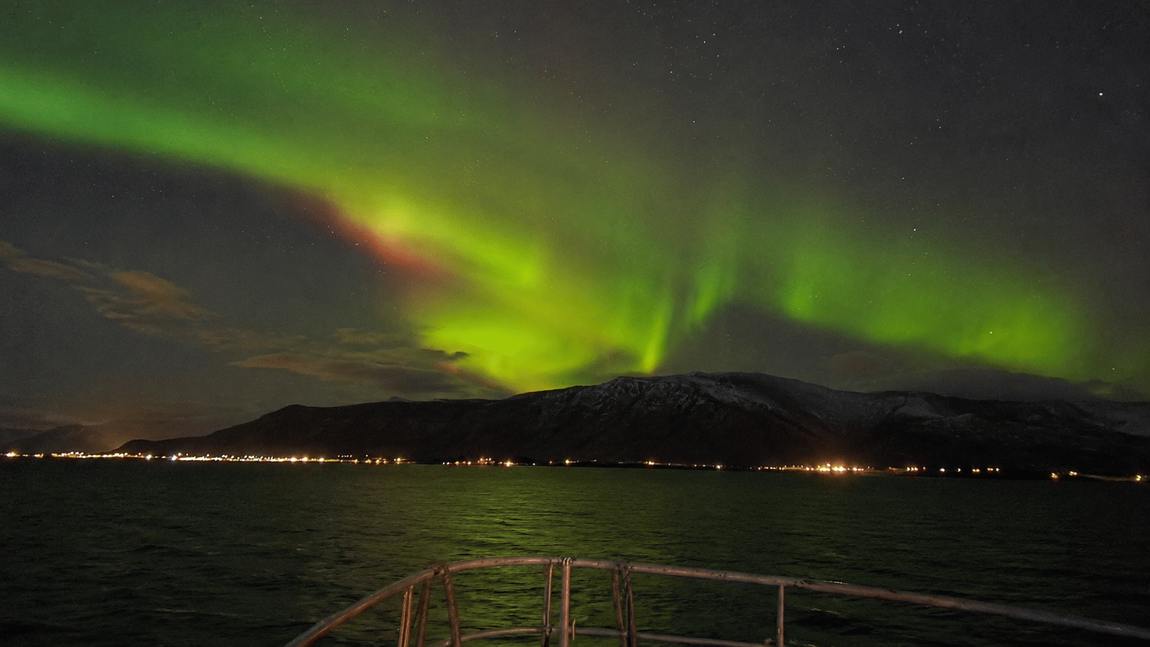 Green, yellow and pink Northern Lights above distant mountains and illuminated coastal buildings.
