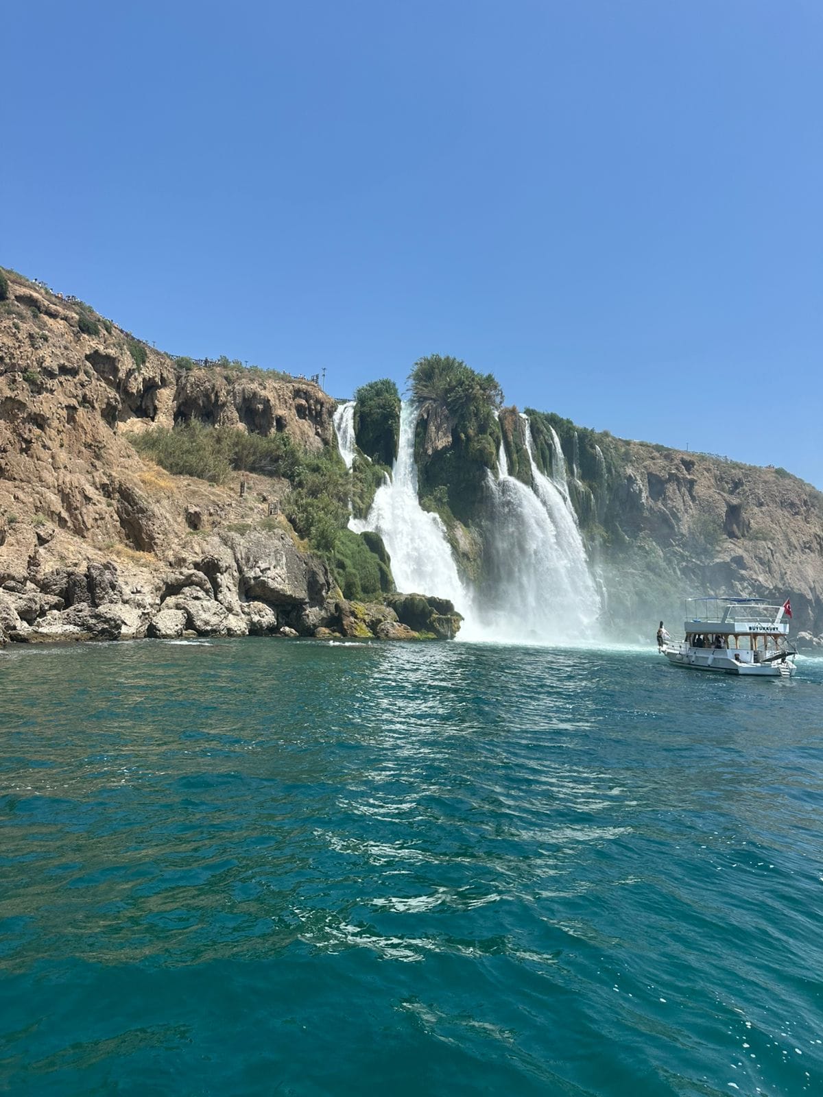 “Our tour group on a boat cruising near the cascading Duden Waterfall in Antalya, surrounded by lush greenery and clear waters.”