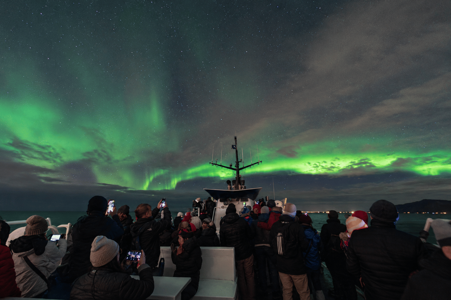Group of passengers watching green aurora borealis from an outdoor viewing deck at night.
