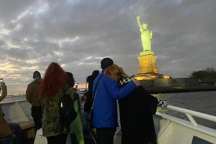 Tourists on a ferry viewing the illuminated Statue of Liberty at sunset over New York Harbor