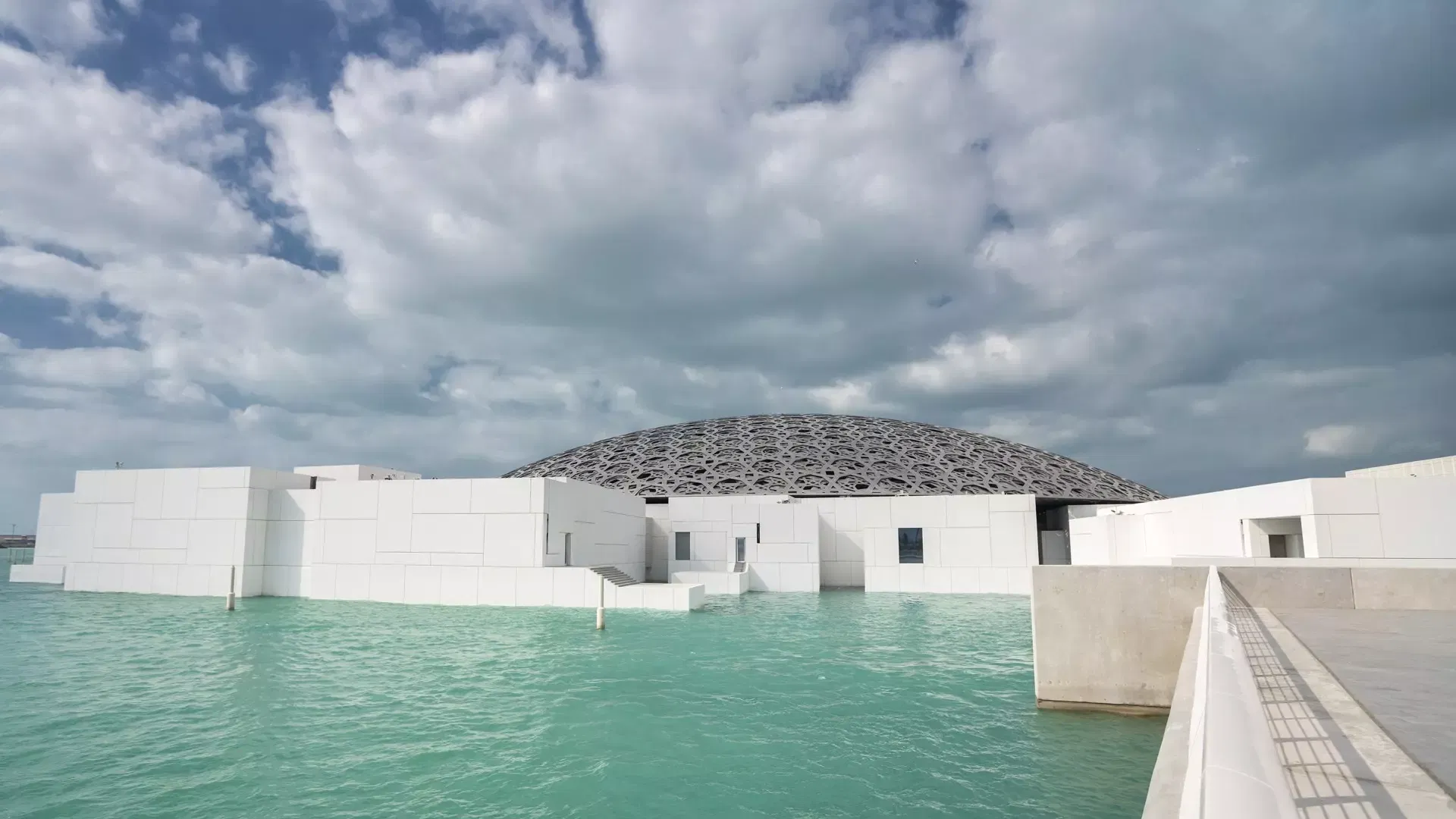White cubic gallery buildings standing in bright green shallow water under a heavy cloudy sky, with a walkway leading in, at Louvre Abu Dhabi