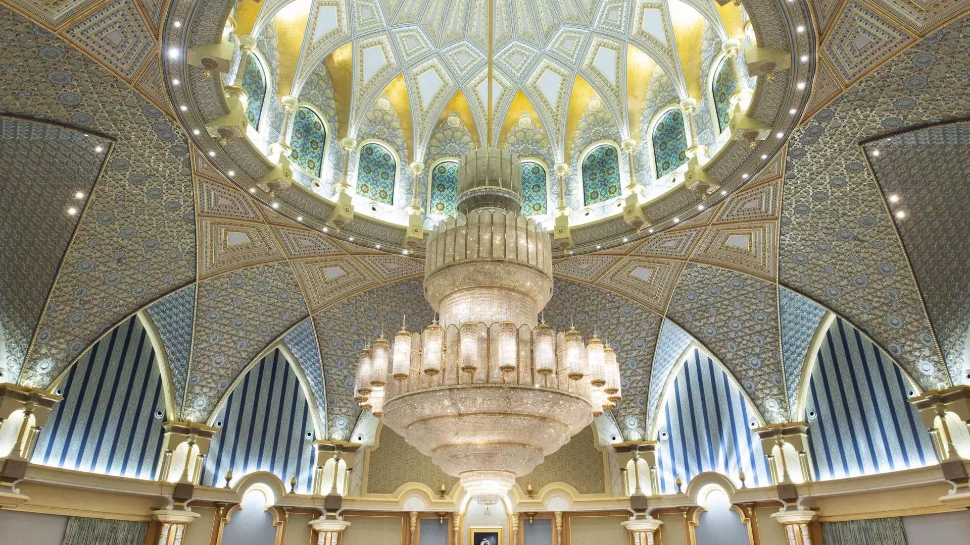 A low-angle interior view showcasing a massive tiered crystal chandelier hanging beneath a soaring domed ceiling adorned with gold trim, stained glass windows, and blue-and-white patterned vaults at Qasr Al Watan, Royal Palace.