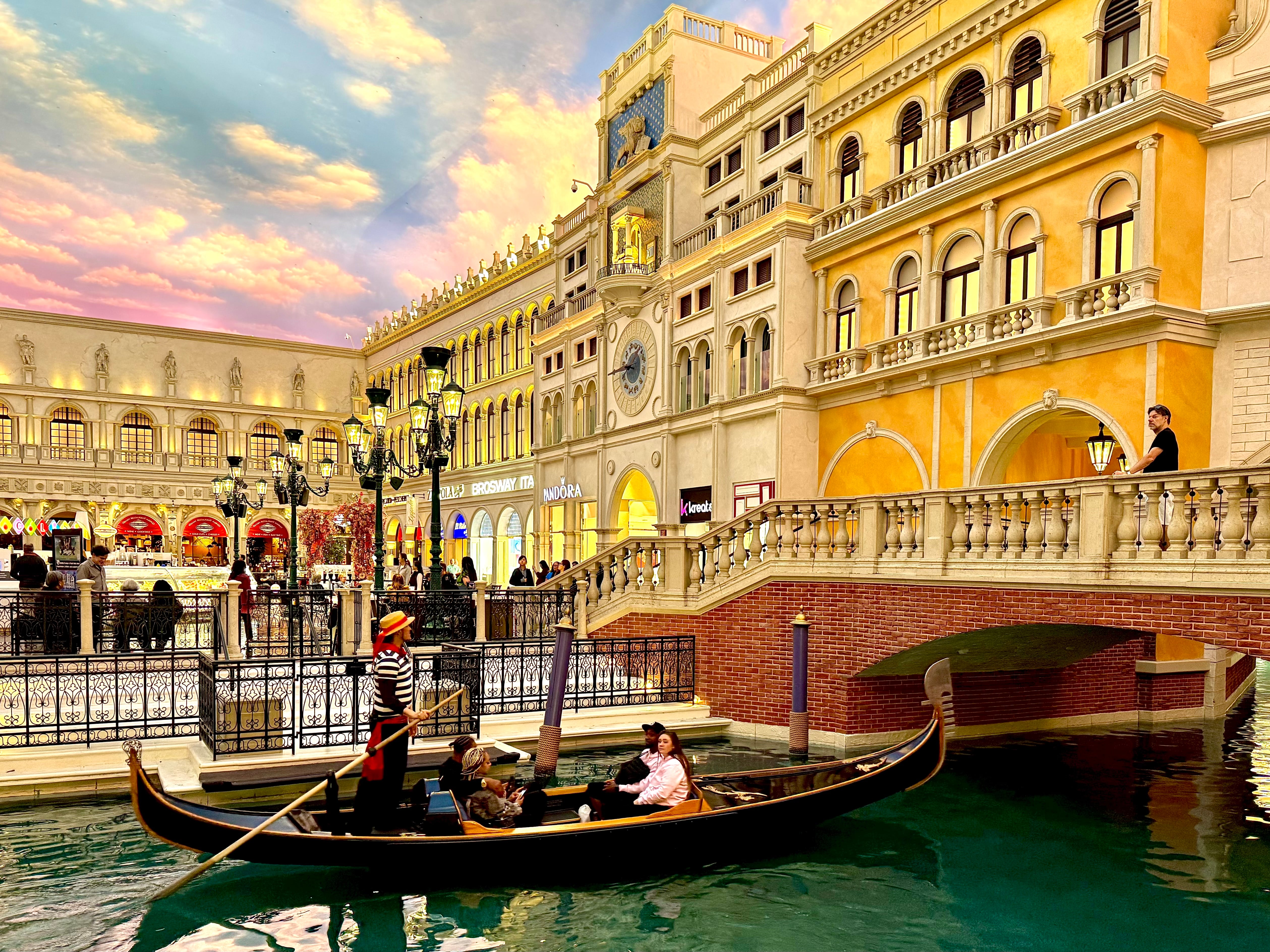 An indoor canal with gondolas surrounded by Venetian-style buildings beneath a painted blue sky.