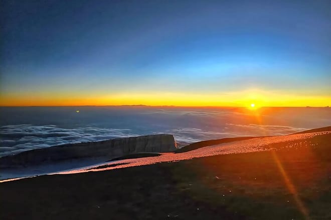 The sun rising over a sea of clouds from a snowy mountain peak, with orange and blue hues in the sky.