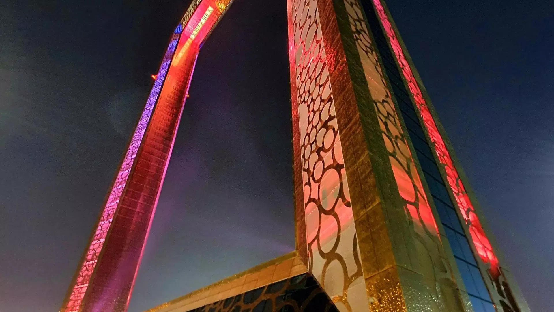 A low-angle night view of one corner of the structure, the gold cladding washed in pink and red light against a dark sky at Dubai Frame