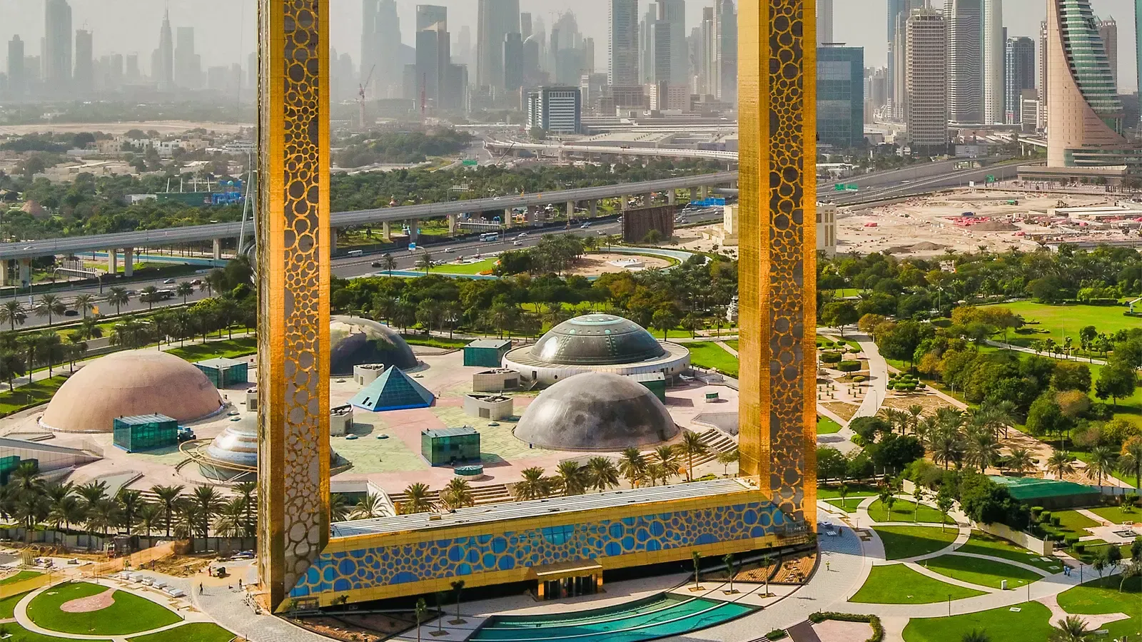An elevated daytime view between the two golden legs of the structure, looking down on the park's domed pavilions and out to the city towers at Dubai Frame