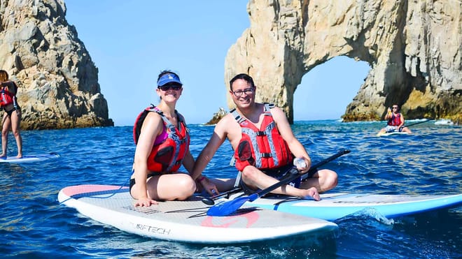 Couple paddle boarding near the Arch of Cabo San Lucas wearing life jackets on calm ocean water