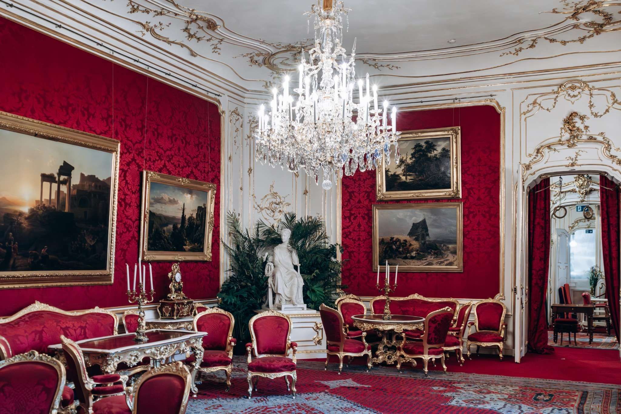 Ornate room with red walls, gold-trimmed furniture, chandeliers, and historic paintings inside the Sisi Museum