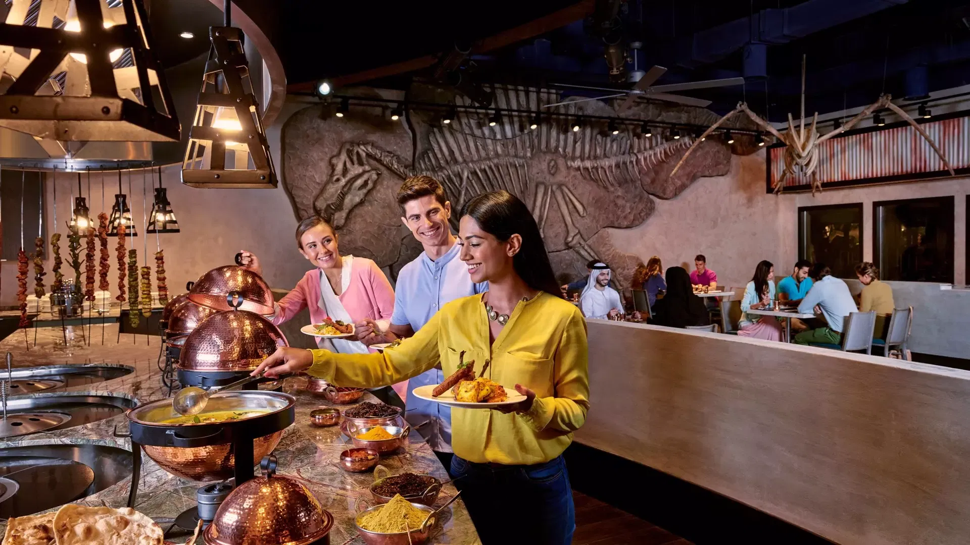 Guests select dishes at a buffet line featuring copper serving bowls while others dine beneath a wall art mural of a dinosaur skeleton at IMG Worlds of Adventure Theme Park.