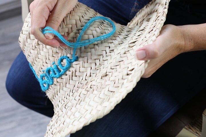 Close-up of a hand embroidering a word with bright blue thread on a straw hat during the personalization workshop.