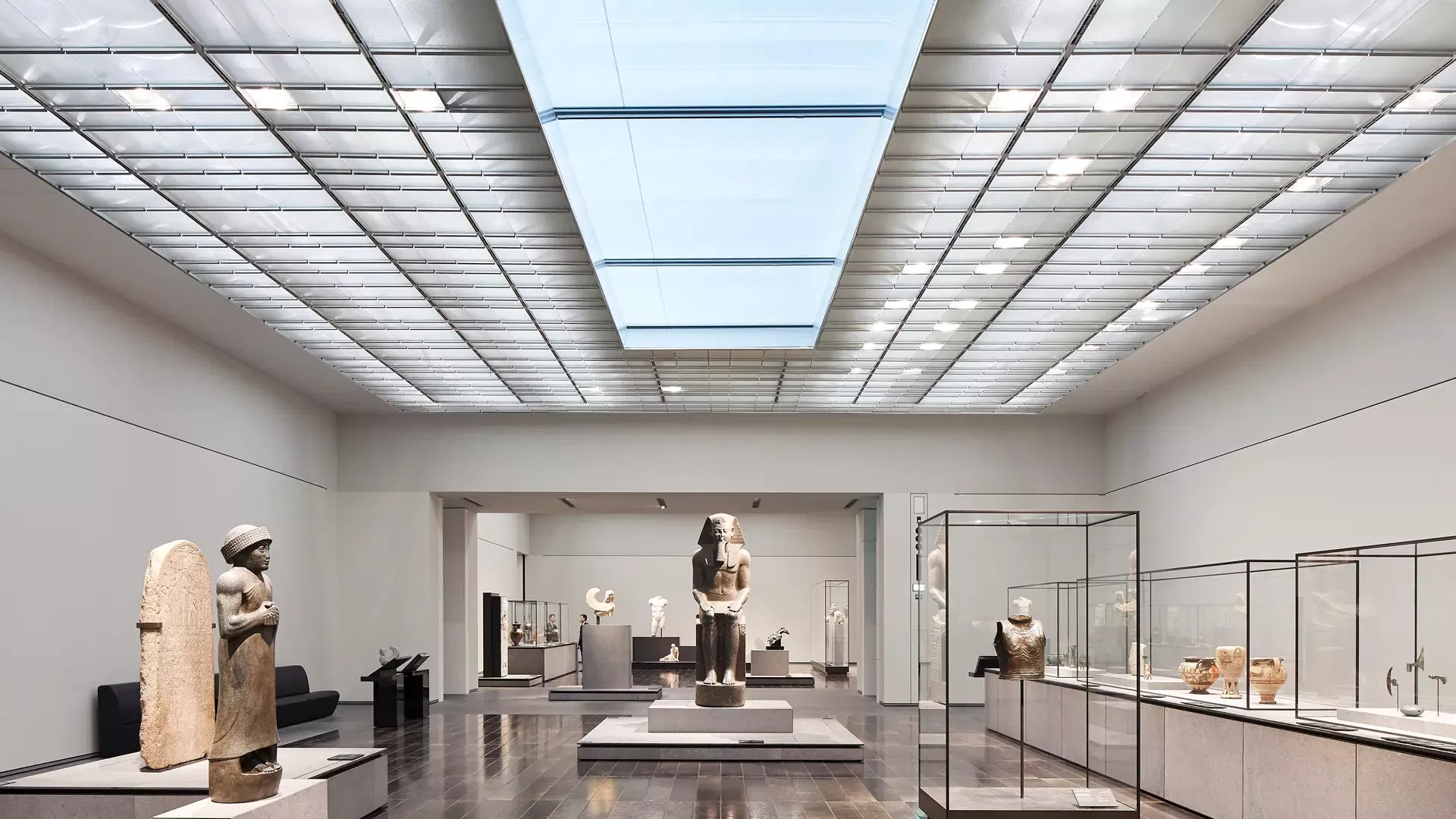 A daylit gallery of ancient stone statues, a seated pharaonic figure and glass display cases of antiquities beneath a skylight at Louvre Abu Dhabi