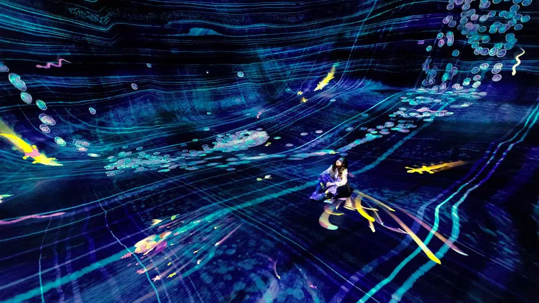 A visitor sits on the floor of a vast, curving blue digital environment filled with swirling glowing lines and shapes at TeamLab Phenomena.