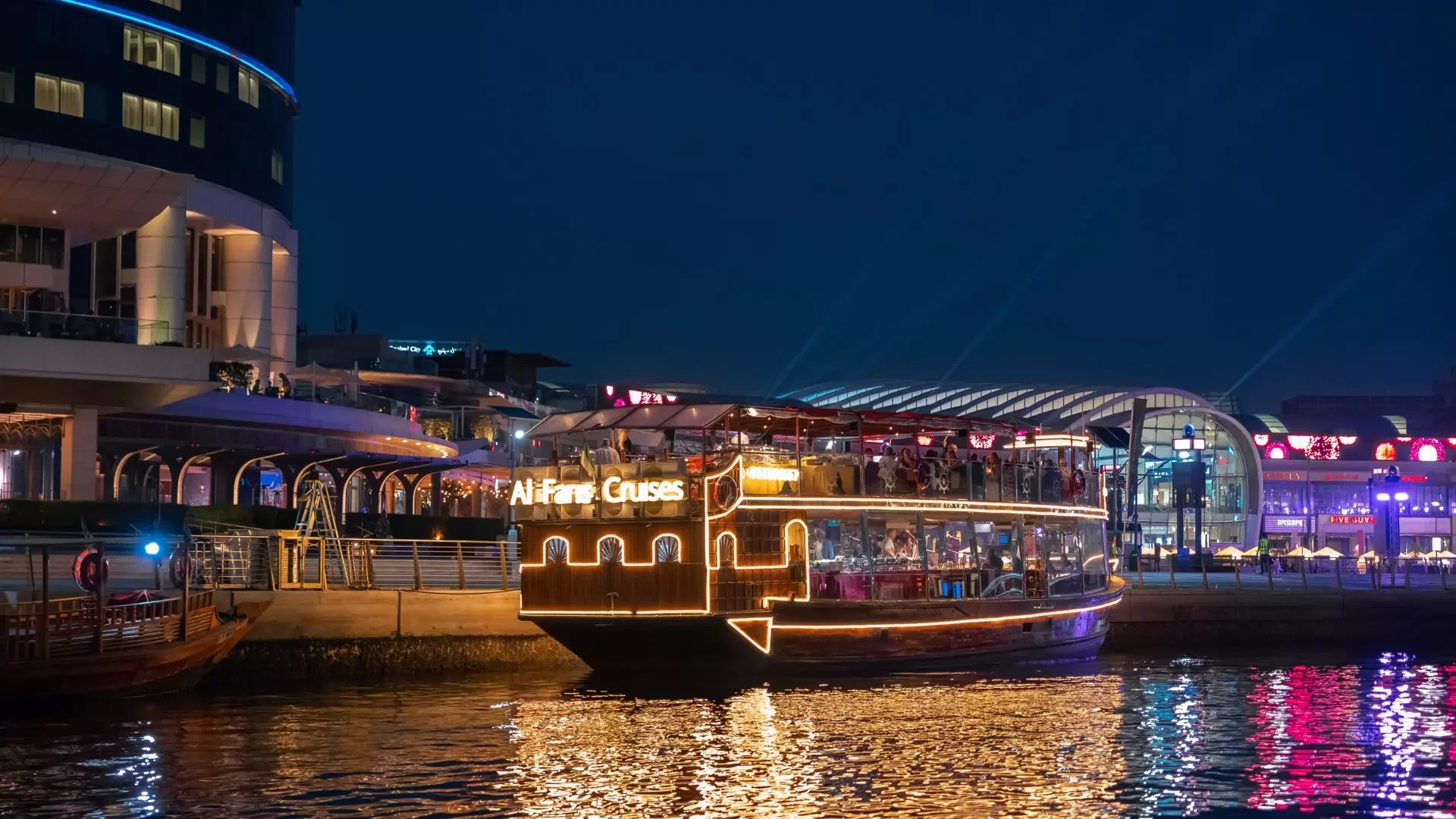 A large, illuminated tourist boat with lights reflecting on the dark water is docked alongside a modern waterfront at Dubai Water Canal Dhow Cruise.