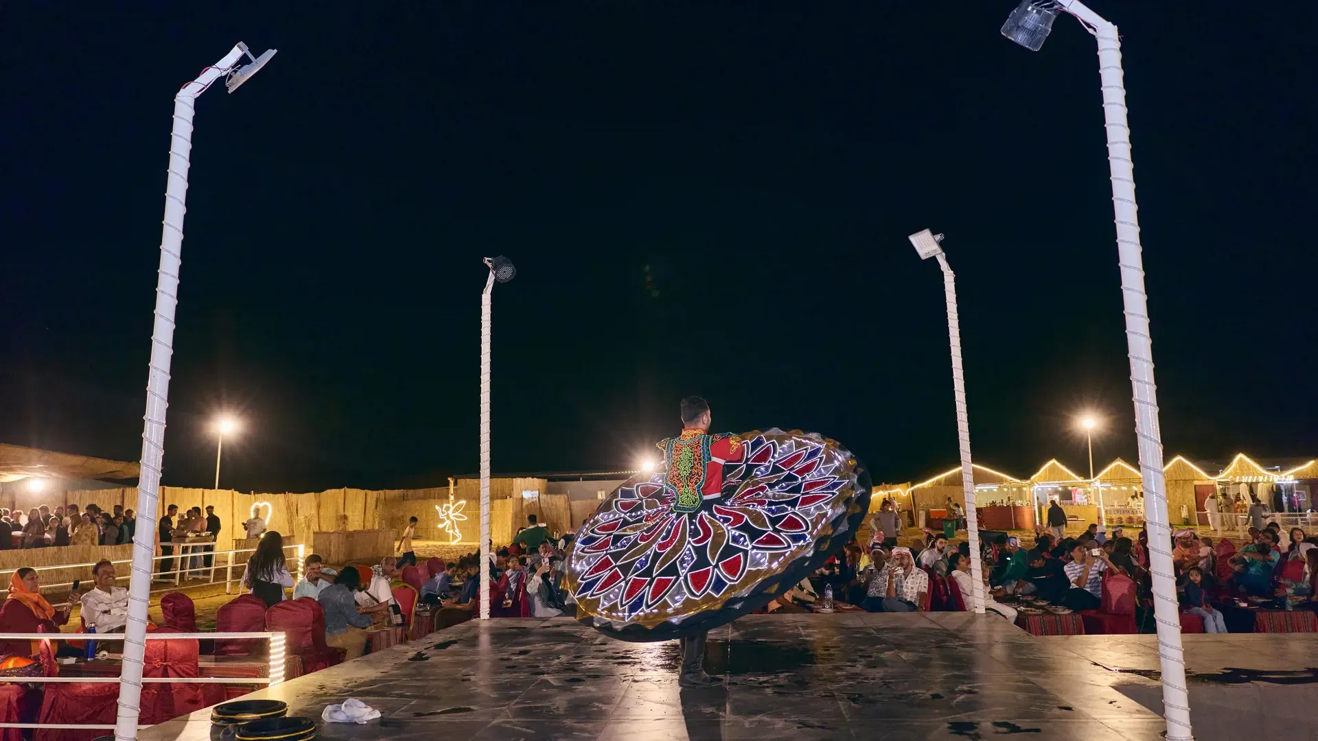 A performer in a vibrant, patterned skirt spins on an outdoor stage at night at Desert Safari + Al Faqaa Camp.