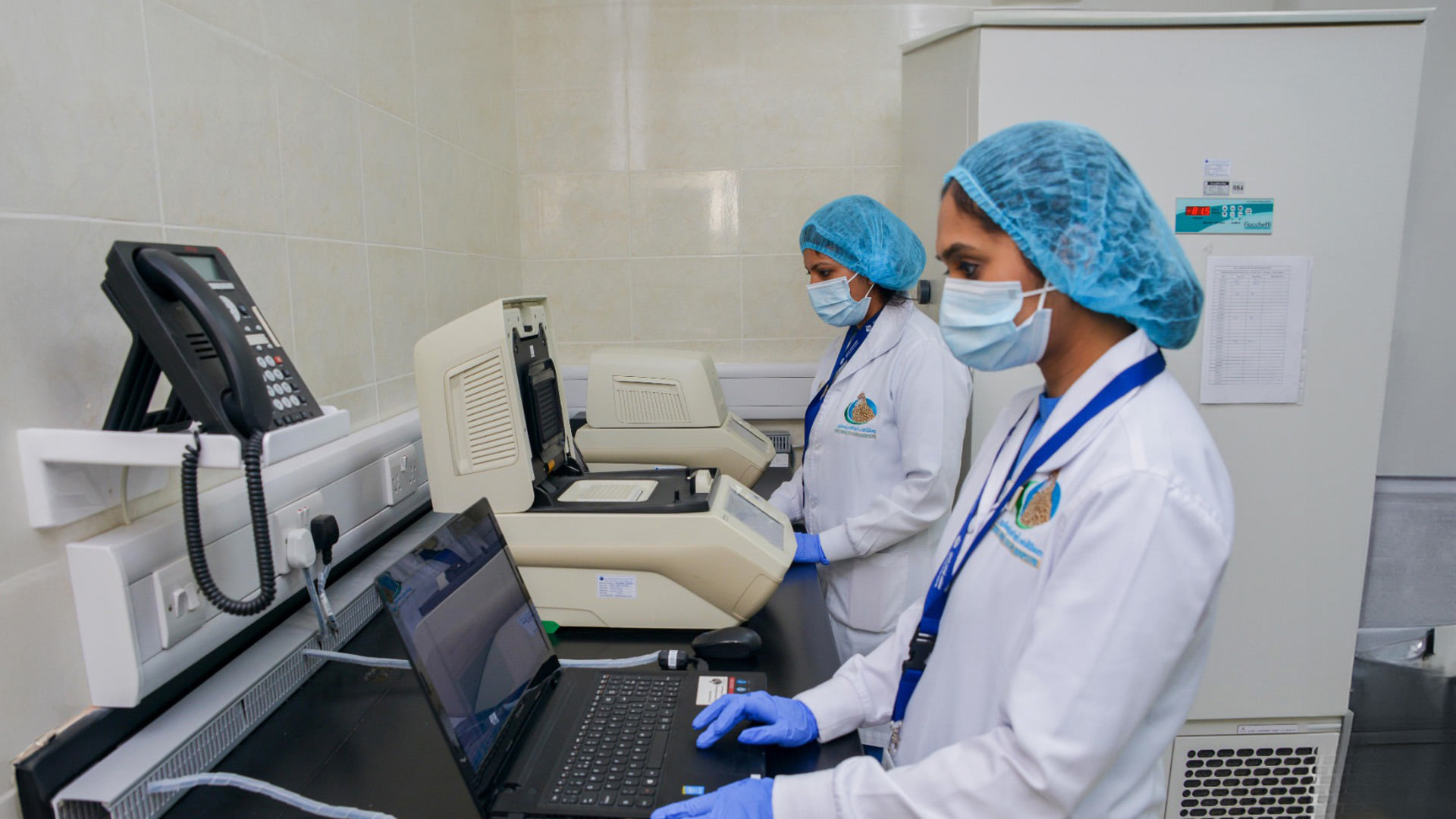 Two lab technicians in white coats, blue hairnets, and face masks operate computers and laboratory equipment at Abu Dhabi Falcon Hospital.