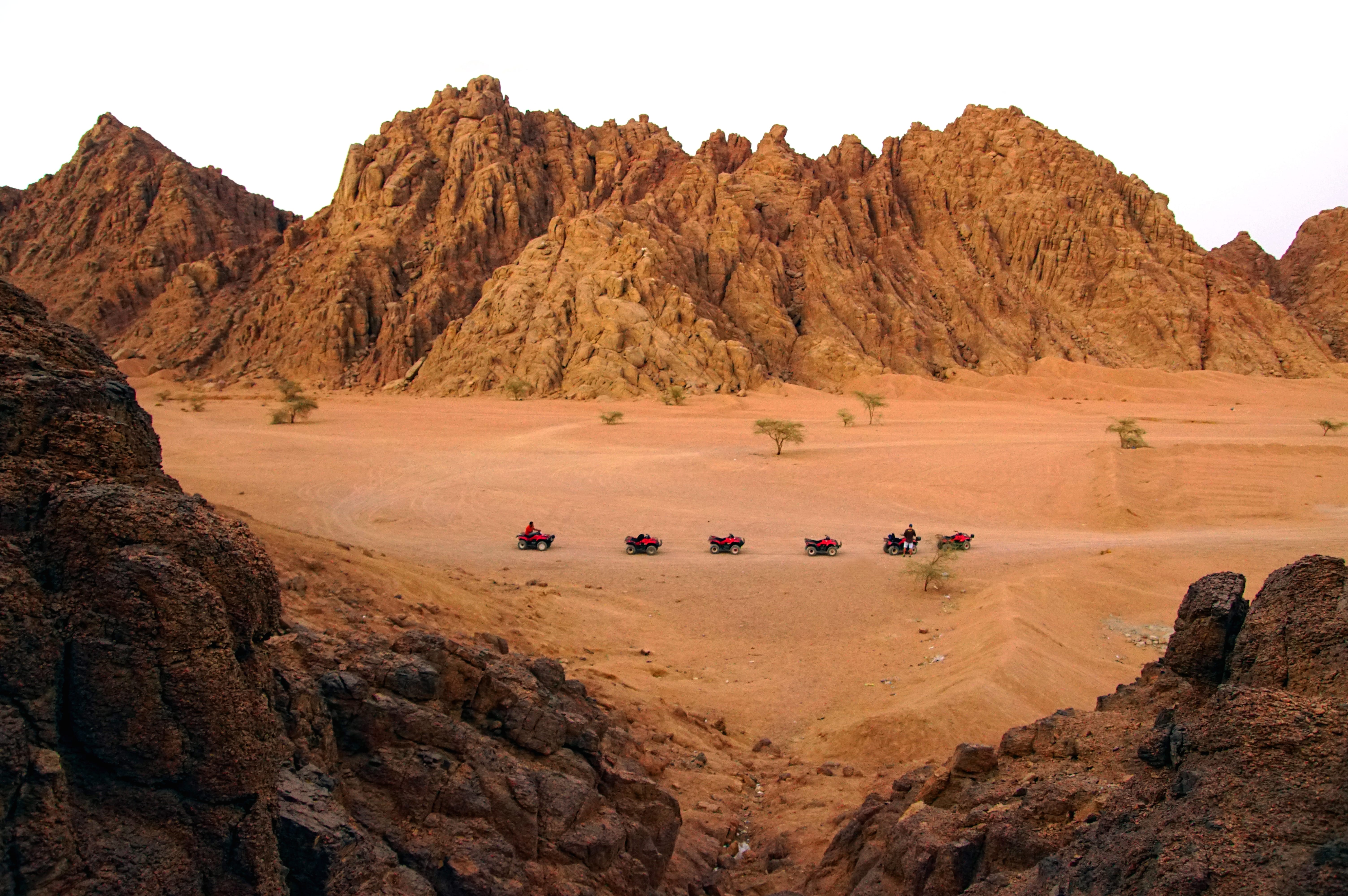 Five quad bikes driving in a line across the flat, sandy desert floor framed by large, rugged, brown rock formations and mountains.