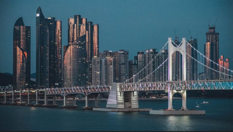 Gwangandaegyo Bridge illuminated at dusk, with Busan's skyline of high-rise buildings glowing in the background.
