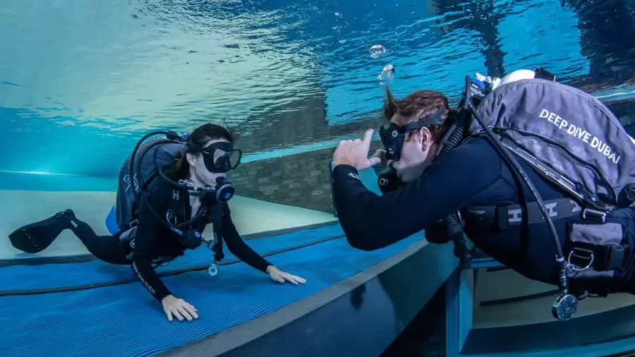Two scuba divers face each other underwater, communicating near a blue platform at Deep Dive Dubai.