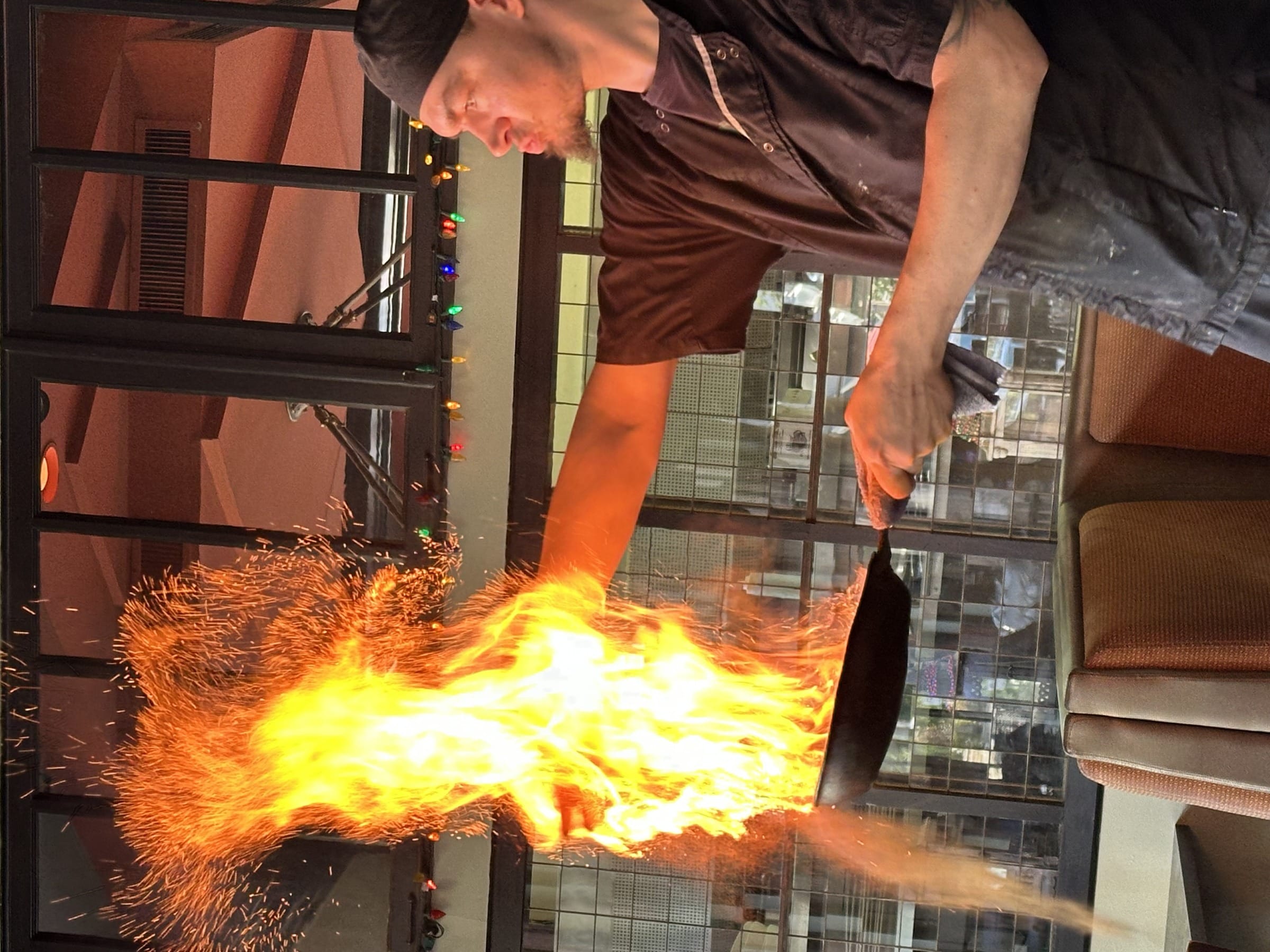Chef tossing a pan with dramatic flames inside a restaurant, dark interior with string lights