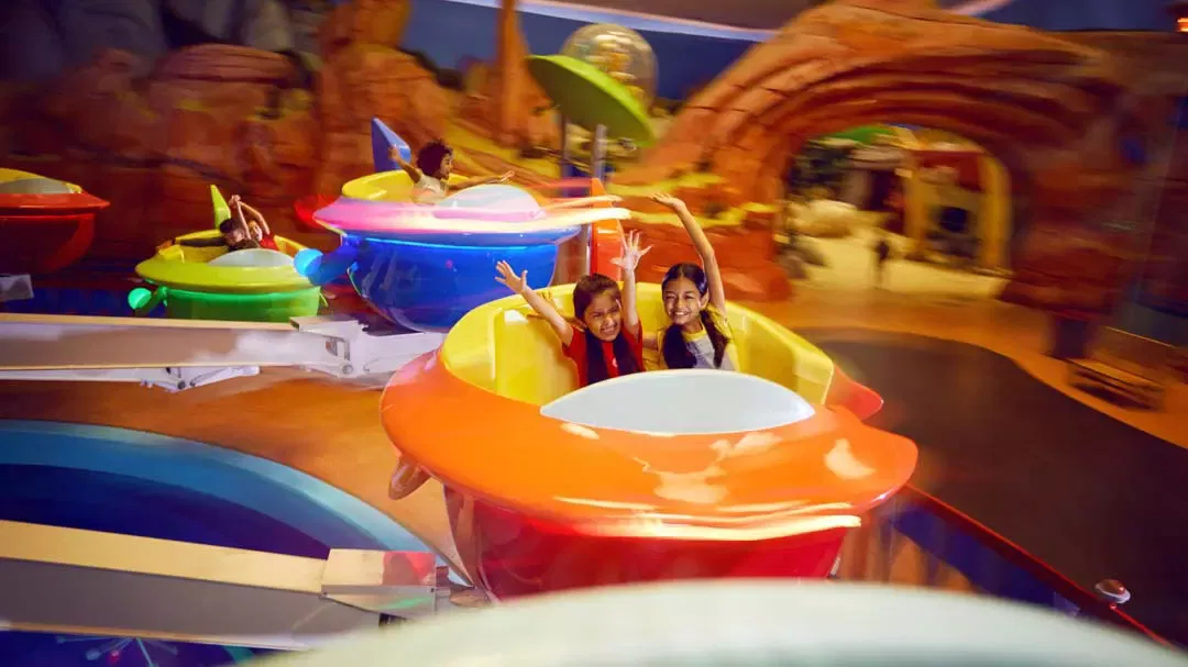 Two girls riding in a bright orange spinning-cup ride past rocky archway scenery at Warner Bros. World Abu Dhabi Theme Park.