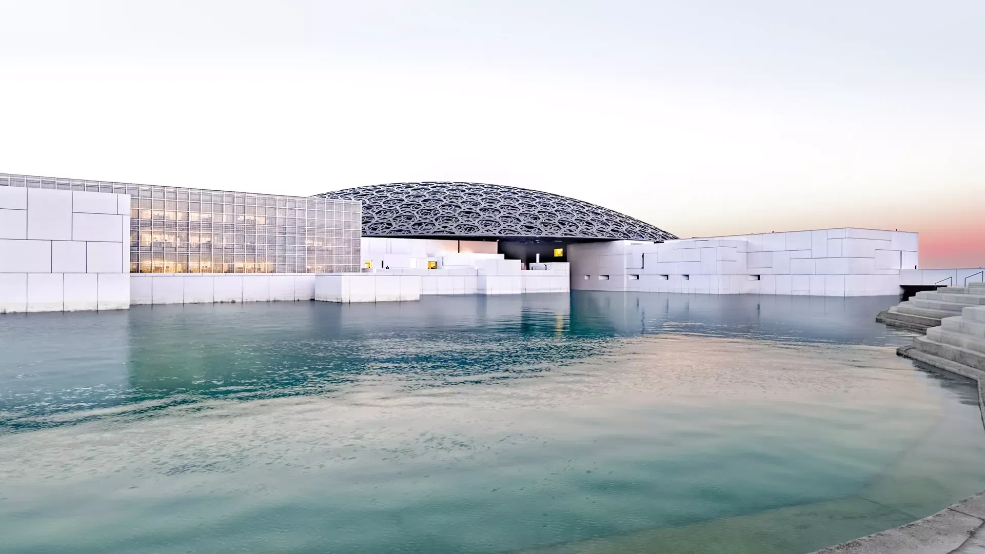 The museum seen across still water at dusk, the lit glass facade and the flattened dome mirrored in a pink and blue sky at Louvre Abu Dhabi