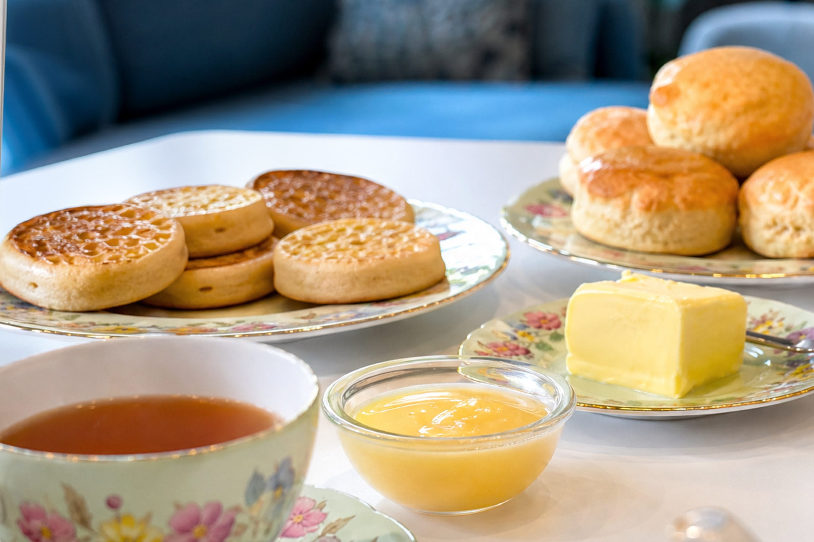 A table laden with baked goods; scones, crumpets, butter, lemon curd and a cup of tea in the foreground