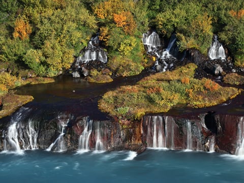 Aerial view of Hraunfossar waterfalls in autumn, with multiple cascades emerging from rust-colored and black lava rock into a vivid turquois