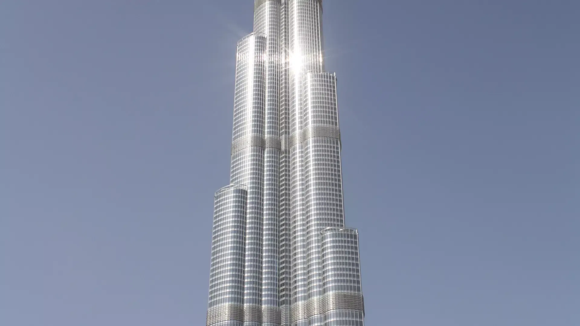 The upper tiers of the tower's silver glass facade catching a burst of sunlight against a pale blue sky at Burj Khalifa